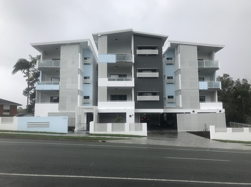 View from the street of a 4-storey development with secure carparking underneath. The windows and balconies include white aluminium screening. The building is painted an off-white colour, with light blue accents.